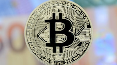 I förgrunden ett mynt med tecknet för bitcoin. i bakgrunden utsuddade eurosedlar.