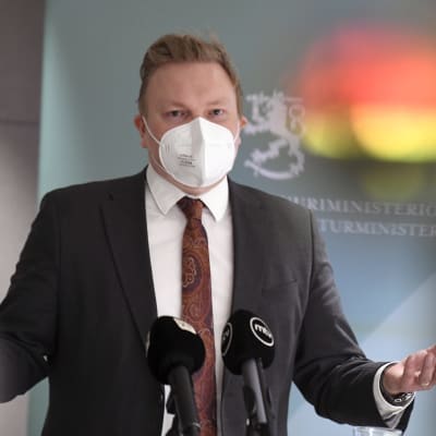 Antti Kurvinen under ett presstillfälle. Han bär ett vitt munskydd.
