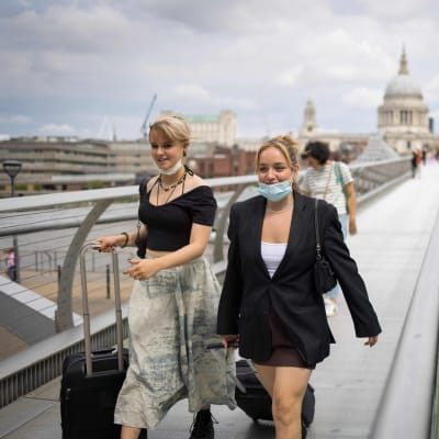 Två unga kvinnor går på Millennium Bridge i London. De bär munskydd under sina hakor.