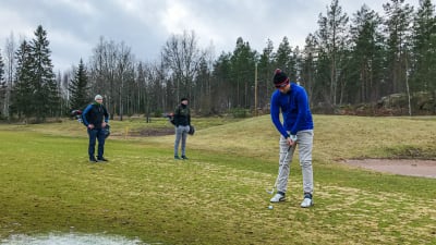 Tre män står på en golfbana, en puttar medan de andra ser på.