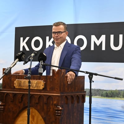 Petteri Orpo står och håller tal vid partimötet i Seinäjoki, bakom sig har han ett sjölandskap. 