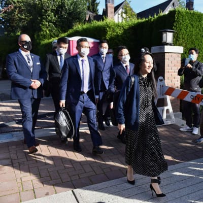 Kvinna i klänning går utomhus i soligt väder med en grupp kostymklädda män i munskydd. Huaweis finanschef Meng Wanzhou lämnar sitt hem i Vancouver den 24 september 2021. 
