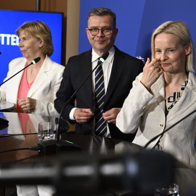 Fyra leende partiordförande i kavajer och kostym på en presskonferens. De är Svenska folkpartiets Anna-Maja Henriksson, Samlingspartiets Petteri Orpo, Sannfinländarnas Riikka Purra och Kristdemokraternas Sari Essayah.