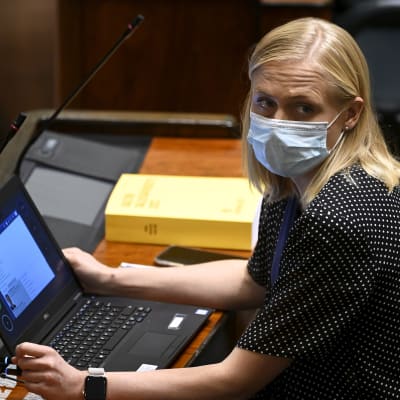 Riksdagsledamoten Elina Valtonen i munskydd sitter i riksdagen med en bärbar dator på bordet hon sitter vid. 
