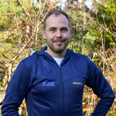 En man med skägg står i solen och ler. Han har en blå jacka med svenska landslagets tryck på.