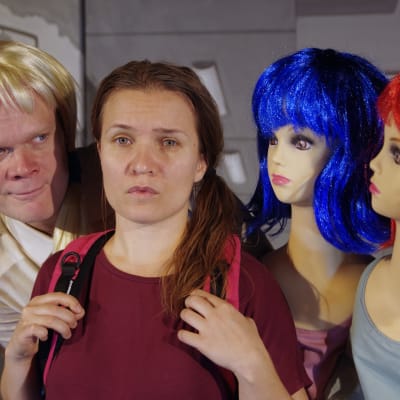 En kvinna ser in i kameran med ledsen blick. Två dockor och en manlig skådis i peruk omringar henne.