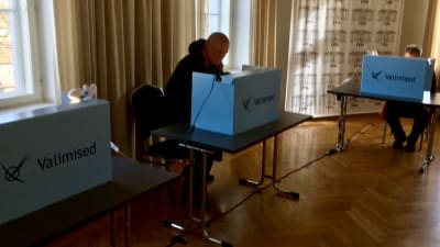 Parlamentsval i Estland 2019 i en mörk sal där två män som bäst håller på att rösta i båset.