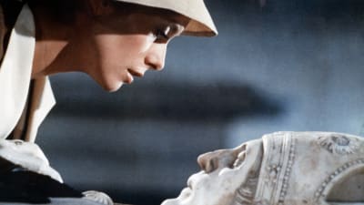 Hattupäinen nainen (näyttelijä Catherine Deneuve) kumartuneena marmoriseen hautapaateen veistetyn piispan muotokuvan ylle. Kuva elokuvasta Tristana.