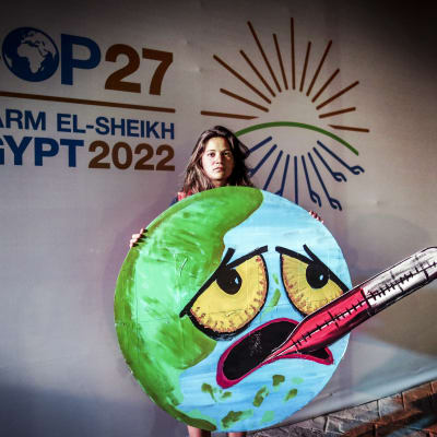 En klimataktivist håller i ett plakat som föreställer ett jordklot med ögon och mun. Ögonen är ledsna och munnen pekar nedåt med en temperaturmätare i. 
