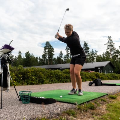 En kvinna står med klubban i luften, beredd att slå till en golfboll.