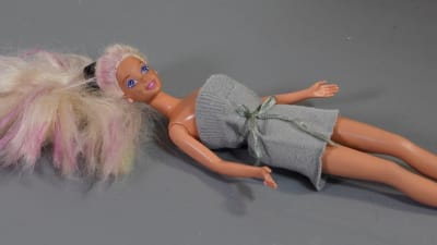 En Barbiedocka ligger på ett bord. Hon har på sig en fodralklänning gjord av en skaftet från en gammal strumpa