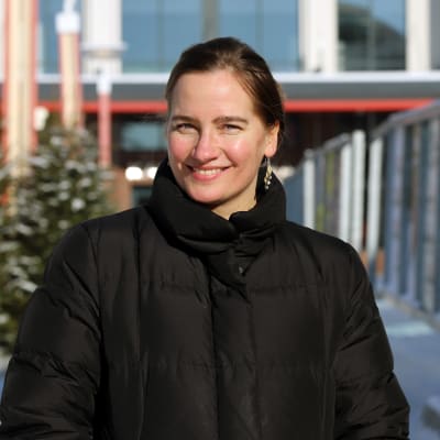 Ruskeahiuksinen nainen katsoo kameraan hymyille, taustalla vaalimainoksia talvisessa kaupunkimaisemassa.
