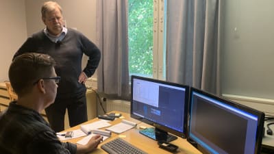 En man står intill ett bord där en kvinna sitter och jobbar vid en dator. 
