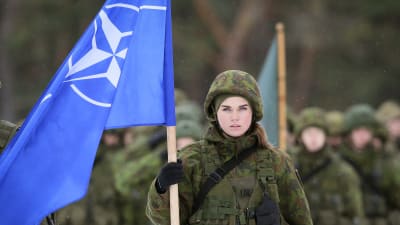En soldat klädd i militärutrustning håller i Natos blåa flagga. I bakgrunden fler soldater.