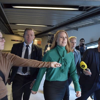 En kvinna marscherar bestämmt genom en korridor med säkerhetsteam och journalister som sticker fram mikrofoner.