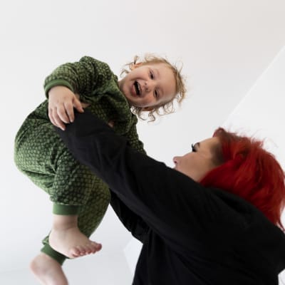 Punahiuksinen nainen pitelee parivuotiasta nauravaa lasta korkealla ilmassa käsillään.