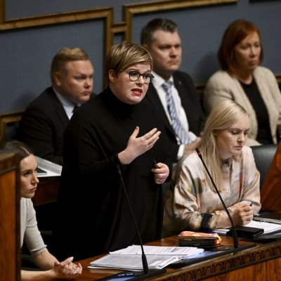 Annika Saarikko puhuu eduskunnassa, ympärillä muita ministereitä