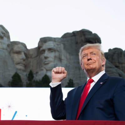 Trump i kostym gör en knytnävsgest med ena handen framför Mount Rushmore.
