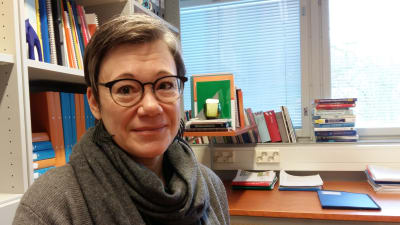 Niina Junttila, en dam med glasögon och grånande hår och grå tröja, sitter i sitt arbetsrum med böcker, skrivbord och en spjälgardin.