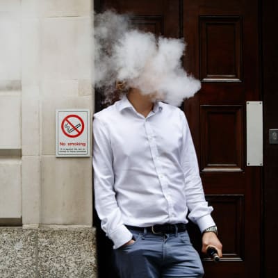 En man står framför en vägg och röker en e-cigarett. Huvudet är täckt i ett rökmoln.