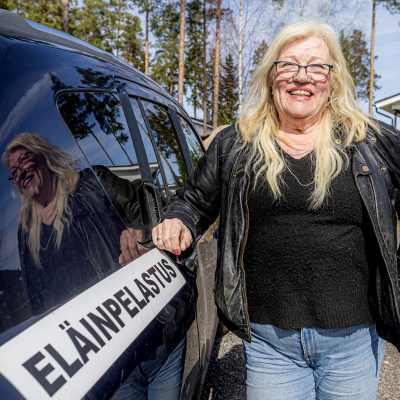 En kvinna, Britt-Marie Juup, står bredvid en bil där det står "eläinpelastus". På svenska betyder det djurräddning. Hon lutar sig mot bilen med ena armen. I bakgrunden syns en uppfart till ett hus.