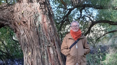 Litteraturforskaren Rita Paqvalén med stor orange halsduk i skogen intill en tjock trädstam. 2021