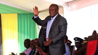 En äldre svart man i kostym står och vinkar med handen. Bakom honom sitter fler personer. John Magufuli inledde en ny presidentperiod i november 2020.