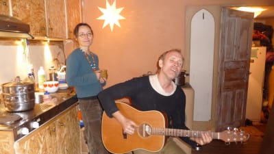 Charlie Jespergaard  spelar gitarr och sjunger. Bakom honom står Petra Blomqvist med en temugg i handen. De är hemma