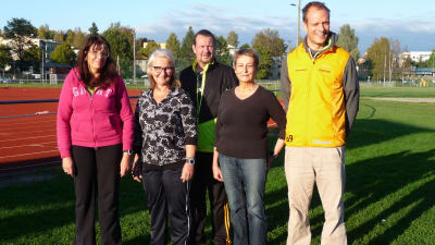 bild på motionärerna Synnöve Lindholm, Mirja Koponen och hennes man Arto, Karin Svahhnström och Heikki Kiili. De poserar ute på Karis idrottsplan med löpbanorna i bakgrunden. Solig kväll i september.
