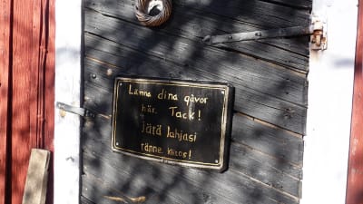 en svart skylt på en svartmålad trädörr. På skylten står det att man kan lämna sina gåvor där, på svenska och finska.