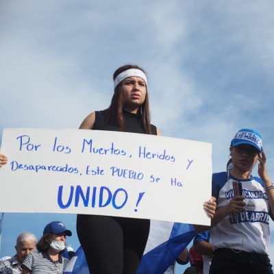 En ung kvinna med pannband håller upp en skylt med texten Por los Muertos, Heridos y Desaparceidos, este Pueblo se ha Unido!