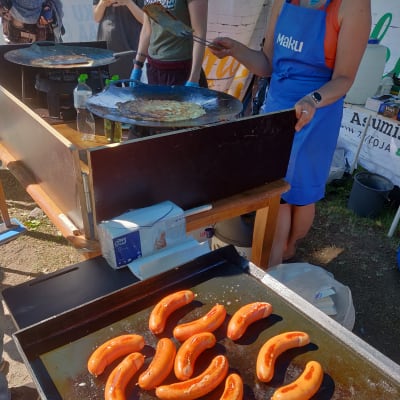 På en grill ligger grillkorv och steks. I bakgrunden ser man händer som steker plättar på en panna.