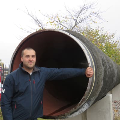 En man står vid ett cirka tolv meter långt stålrör beklätt av betong. Diametern är drygt en meter. Röret finns i Hangö, liggande på cementpelare.
