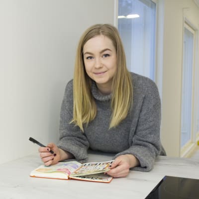 Caroline Eriksson har långt blont hår och en grå tröja. Hon lutar sig över ett bord med ett häfte framför sig. I handen håller hon en penna. 