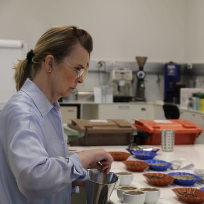 En kvinna klädd i ljusblå skjorta arbetar i ett laboratorium. På bordet framför henne står ett antal skålar med kaffebönor och kaffe i vita koppar.  