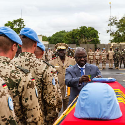 Stupade FN-soldater hedras på flygfältet i Bamako, Mali. En blå fredsbevararhjälm vilar på en kista som är täckt med tyska flaggan. En svart man i kostym lägger en medalj på kistan medan soldater i blå baskrar står i rad intill.