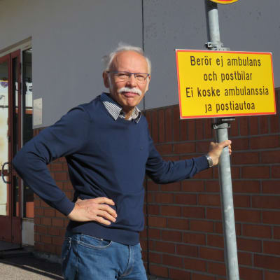 En man med grått hår, mustasch och blå tröja står utanför Ingå hälsocentrals ingång. Han står och håller i en trafikskylt som förbjuder trafik, men med tillägget "berör ej ambulans och postbil".