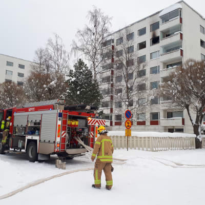 Brand i ett höghus i Karleby, på bilden syns en brandman och en brandbil på gatan framför ett höghus. Högt uppe i höghuset syns ett söndrat och sotat fönster.