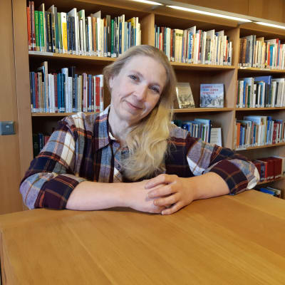 medelålders kvinna med långt blont hår står leende och knyter sina händer framför sig på ljusbrun hylla i bibliotek