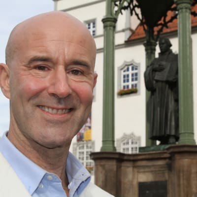 Mark Levengood i vit kavaj poserar på torget i Wittenberg, med staty i Martin Luther i bakgrunden