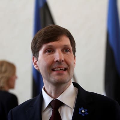 En man står i kostym - i bakgrunden syns Estlands flagga