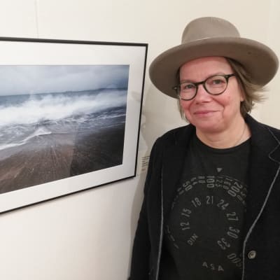 En fotograf står vid sitt havsfotografi "Wave" i ett utställningsgalleri.