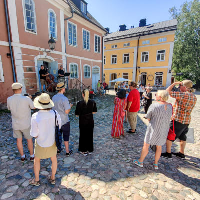Folk står på ett torg och tittar upp på ett hus där en duo spelar musik på en trappa.