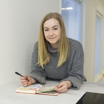 Caroline Eriksson har långt blont hår och en grå tröja. Hon lutar sig över ett bord med ett häfte framför sig. I handen håller hon en penna. 
