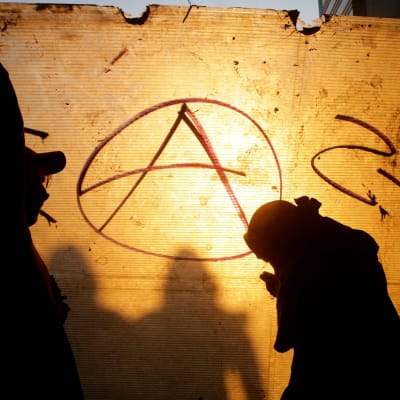 Kolme tummaa hahmoa seinään maalatun anarkistien tunnuksen edessä.