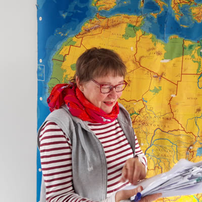 Toimittaja ja ohjaaja Tiina Harpf käsikirjoituspinon kanssa maailmankartan edessä.