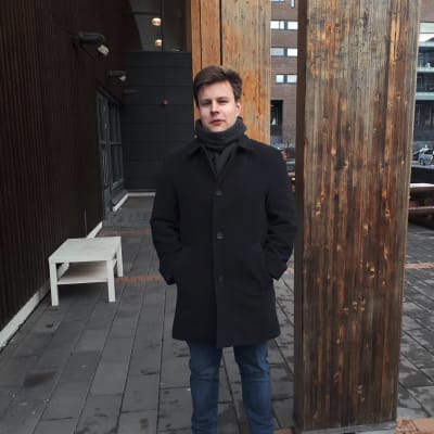 En man iklädd svart jacka står bredvid en mörkare träpelare. 