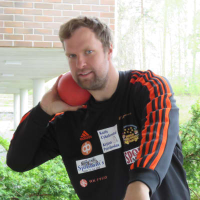 En man med lite lockigt mörkt hår och skägg med en röd kula mot halsen. Han är kulstötare och poserar framför kameran.