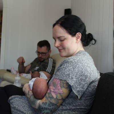 Kvinna som sitter i soffan med ett spädbarn i famnen, i bakgrunden sitter en man med ett annat spädbarn i famnen. 