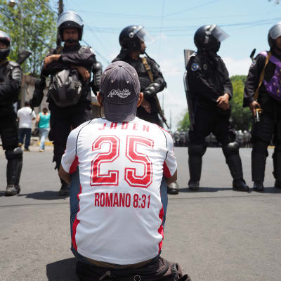Man med vit skjorta sitter på asfalten med ryggen mot kameran, framför står poliser i hjälmar.